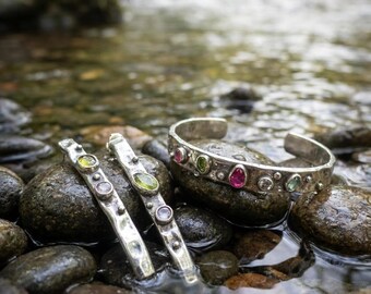 Könnte beinhalten: Zwei Silberarmreifen und ein Silberarmband mit gehämmertem Finish und eingelassenen, mehrfarbigen Edelsteinen in Rosa, Grün und Transparent. Die Schmuckstücke liegen auf nassen Flusskieselsteinen in einem flachen Bachlauf.