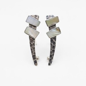 May include: A pair of stud earrings featuring a dark, oxidized silver base with a textured, hammered finish. Each earring displays two rectangular, iridescent white mother-of-pearl inlays set at an angle along the elongated, curved metal stem.