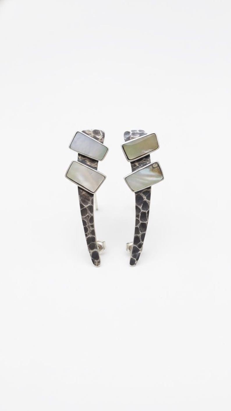 May include: A pair of stud earrings featuring a dark, oxidized silver base with a textured, hammered finish. Each earring displays two rectangular, iridescent white mother-of-pearl inlays set at an angle along the elongated, curved metal stem.