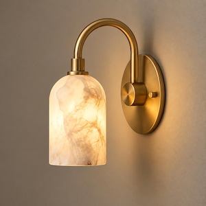 May include: A wall-mounted light fixture featuring a curved brass arm and a circular backplate. The cylindrical shade is made of translucent white material with brown marble-like veining, emitting a warm, soft glow from the internal light source.