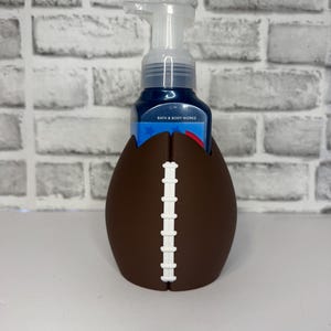 Könnte beinhalten: Ein brauner Seifenspenderhalter aus Silikon in Form eines American Footballs mit weißen Schnürsenkeln, der eine Bath & Body Works Schaumseifenflasche mit einem transparenten Pumpkopf enthält. Der Halter hat eine glatte, matte Oberfläche.