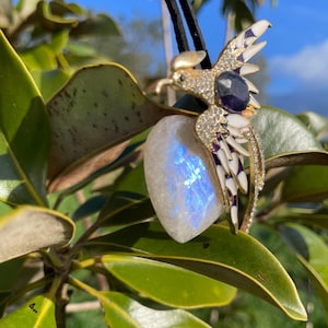 Peut inclure: Pendentif doré représentant un oiseau avec des détails blancs, violets et noirs, orné de strass et d'une pierre précieuse bleu foncé. Il est suspendu à un cordon noir et associé à une pierre de lune irisée en forme de cœur.