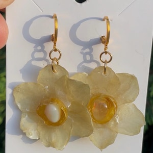 Op de afbeelding: Een paar handgemaakte bloemvormige oorbellen. Elke oorbel heeft een doorschijnende, lichtgele bloem met een gouden ring en een centrale, parelmoer kraal. De oorbellen zijn bevestigd aan een witte kaart met handgeschreven tekst.