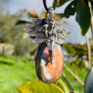 Peut inclure: Un collier pendentif argenté avec une fée ailée tenant un petit objet. Le pendentif est suspendu à un cordon noir et est fixé à une pierre ovale irisée aux teintes roses et orangées. La pierre mesure environ 4 cm de long.