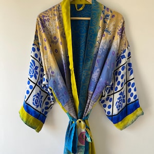 May include: A colorful, patterned robe with a tie closure, hanging on a wooden hanger. The robe features a mix of blue, yellow, and purple hues with floral and geometric designs. The sleeves have a blue and white floral pattern.