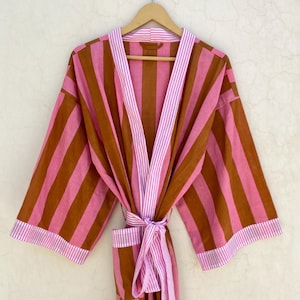 Op de afbeelding: Een gestreepte badjas in roze en bruine tinten, hangend aan een houten hanger. De badjas heeft verticale strepen en een bijpassende riem. De rand en riem zijn geaccentueerd met dunne witte en roze strepen. De badjas is gemaakt van een zachte, lichte stof.