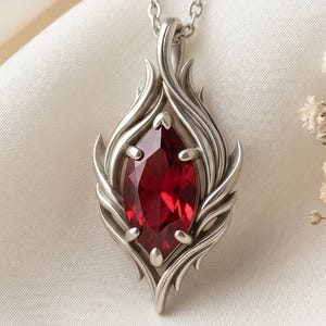 May include: A silver pendant necklace featuring a large, oval-shaped, deep red gemstone. The gemstone is set within a stylized, flame-like silver frame. The necklace is displayed on a white, textured surface.