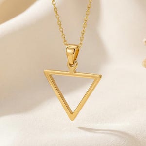 May include: A gold-colored pendant necklace featuring a geometric triangle design. The pendant is suspended from a delicate chain. The necklace is displayed on a white, textured fabric.