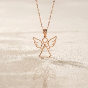 May include: Rose gold angel pendant necklace. The necklace features a delicate angel pendant with outstretched wings and clasped hands. The pendant hangs from a fine chain. The necklace is displayed on a textured, off-white surface.