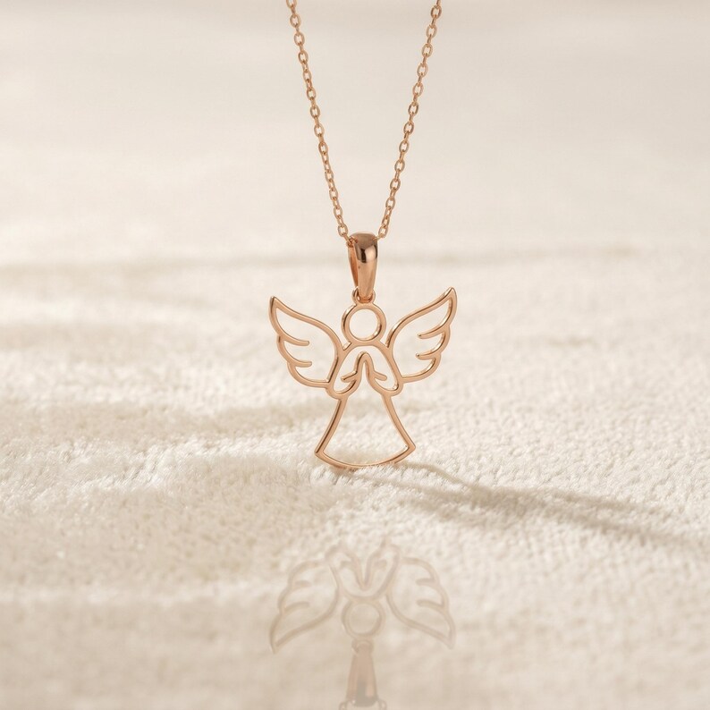May include: Rose gold angel pendant necklace. The necklace features a delicate angel pendant with outstretched wings and clasped hands. The pendant hangs from a fine chain. The necklace is displayed on a textured, off-white surface.