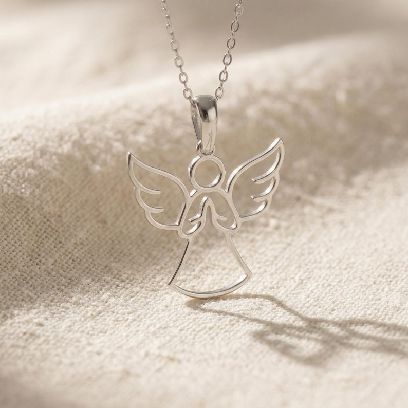 May include: A silver-colored pendant necklace featuring an angel design. The angel has outstretched wings and clasped hands. The necklace hangs on a delicate chain. The pendant is displayed on a textured, off-white surface.