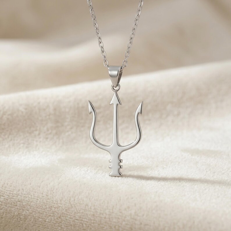 May include: A silver-colored pendant necklace featuring a trident design. The trident has a central arrow-shaped prong flanked by two curved prongs. The necklace hangs on a delicate silver chain, set against a soft, neutral background.