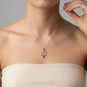 May include: A silver-colored necklace with a trident pendant. The necklace is worn around the neck. The trident pendant is a three-pronged fork design. The necklace is displayed against a neutral background.