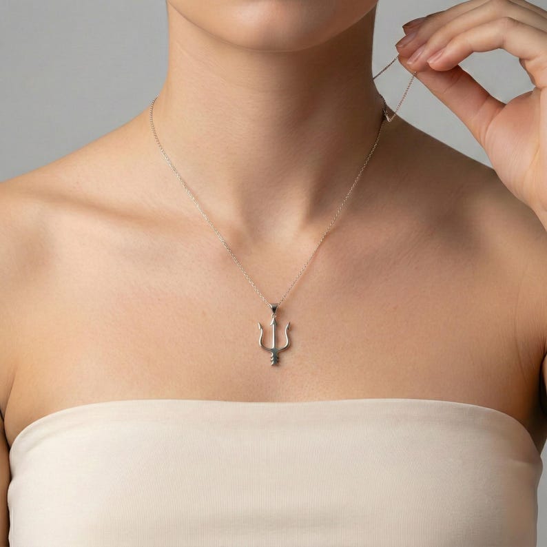 May include: A silver-colored necklace with a trident pendant. The necklace is worn around the neck. The trident pendant is a three-pronged fork design. The necklace is displayed against a neutral background.
