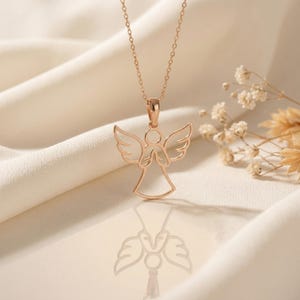 May include: Rose gold angel pendant necklace. The necklace features a delicate, openwork angel design with outstretched wings and clasped hands. The pendant hangs from a fine chain. The background is a soft, neutral color, with a hint of floral accents.