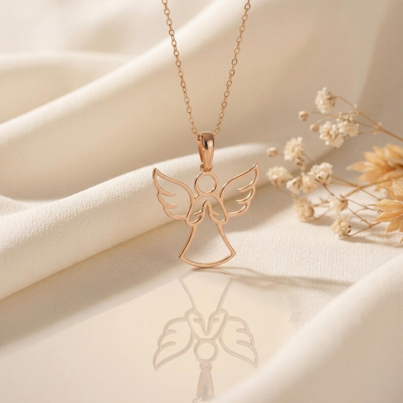 May include: Rose gold angel pendant necklace. The necklace features a delicate, openwork angel design with outstretched wings and clasped hands. The pendant hangs from a fine chain. The background is a soft, neutral color, with a hint of floral accents.