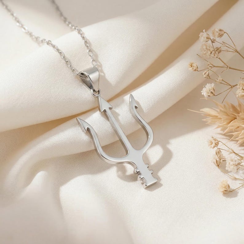 May include: A silver-colored trident pendant necklace. The pendant features a three-pronged spear design with a central arrow-shaped point. The necklace hangs on a delicate chain, displayed on a white fabric background. The trident is a symbol of power and authority.