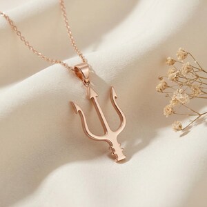 May include: Rose gold trident pendant necklace. The pendant features a three-pronged design with an arrow-shaped center. The necklace is displayed on a white, textured surface with dried flowers in the background.