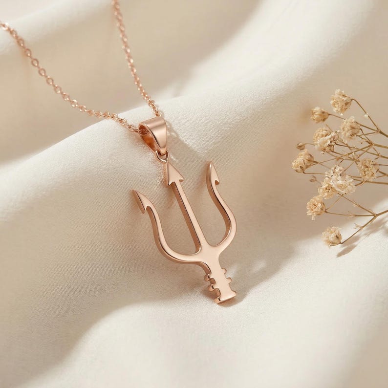 May include: Rose gold trident pendant necklace. The pendant features a three-pronged design with an arrow-shaped center. The necklace is displayed on a white, textured surface with dried flowers in the background.