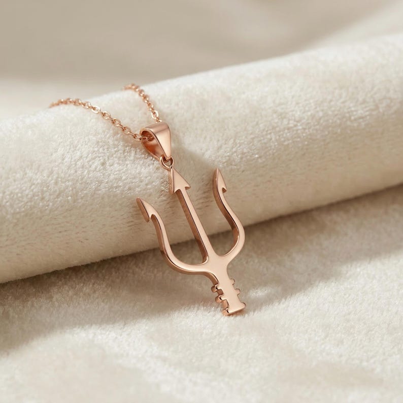 May include: Rose gold trident pendant necklace. The pendant features a three-pronged design with an arrow-shaped center. The necklace is displayed on a soft, white surface.