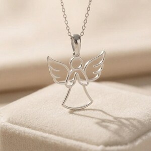 May include: A silver-colored angel pendant necklace. The pendant features an angel with outstretched wings and clasped hands. The necklace is on a delicate chain, and the angel pendant is casting a shadow on the surface below.