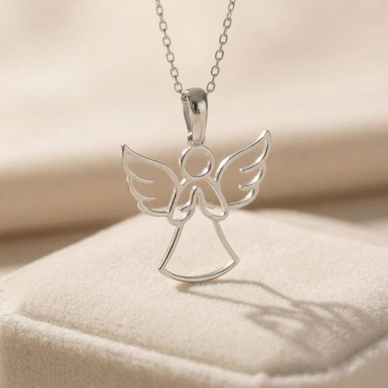 May include: A silver-colored angel pendant necklace. The pendant features an angel with outstretched wings and clasped hands. The necklace is on a delicate chain, and the angel pendant is casting a shadow on the surface below.