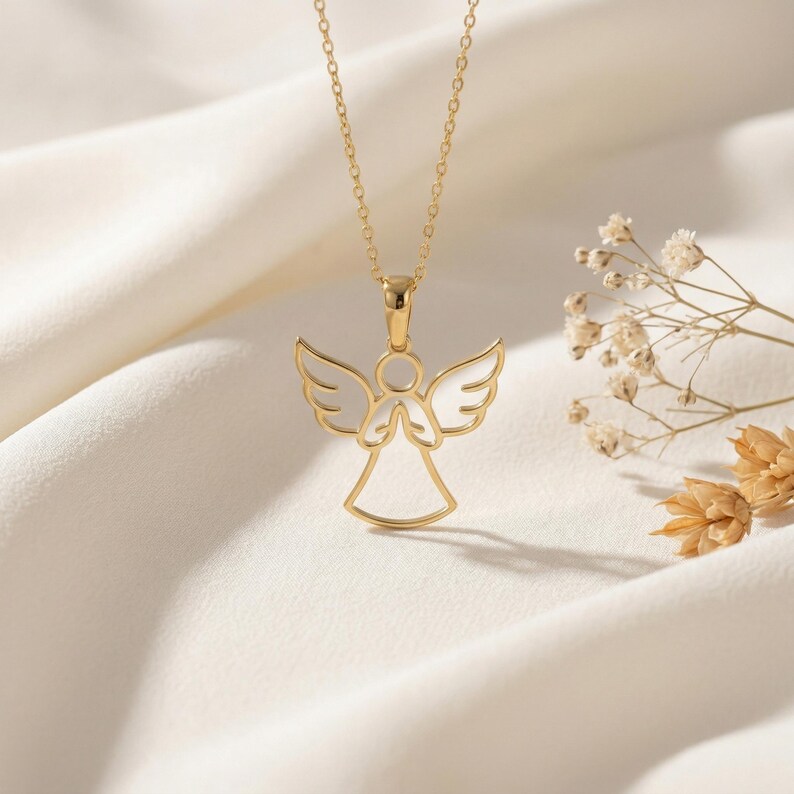 May include: A gold-colored angel pendant necklace. The pendant features an openwork design of an angel with outstretched wings and clasped hands. The necklace is displayed on a white, textured surface.
