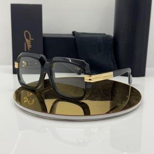May include: Black-framed spectacles with gold-tone metal accents on the temples and hinges. The frames feature a bold, square-shaped design. Included are a black branded case, a black cleaning cloth, and a black display card with the Cazal logo.