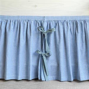 Peut inclure: Un meuble blanc avec un rideau à rayures bleu clair et blanc. Le rideau est noué avec deux nœuds bleu clair. Le meuble est placé sur un fond en bois avec des plantes séchées de chaque côté.