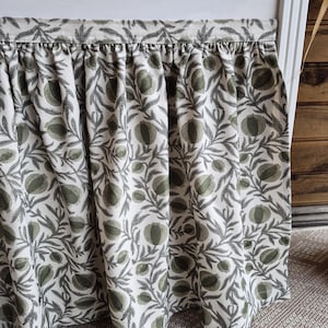 May include: A gathered valance with a botanical print. The fabric displays a repeating pattern of green pomegranates and leaves on a cream backdrop. The valance is attached to a white frame.
