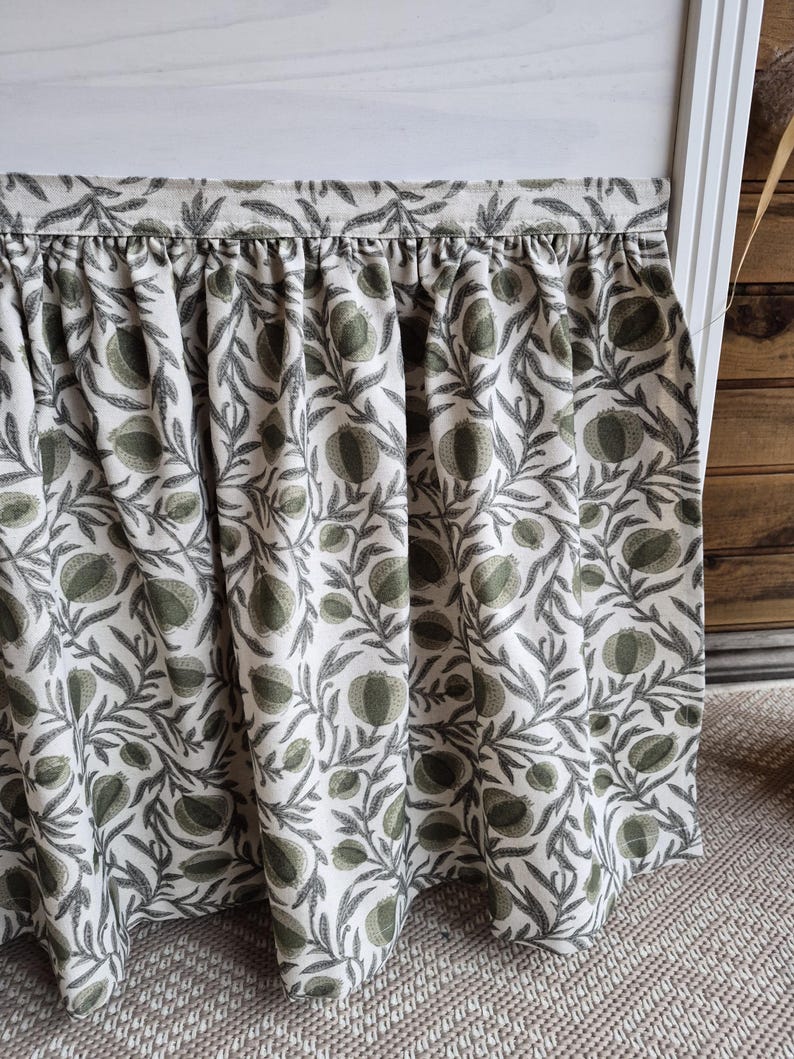 May include: A gathered valance with a botanical print. The fabric displays a repeating pattern of green pomegranates and leaves on a cream backdrop. The valance is attached to a white frame.
