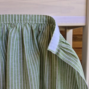May include: A gathered fabric curtain featuring a sage green and white vertical striped pattern. The top edge of the curtain is finished with a white hook-and-loop fastener strip, which is partially attached to a matching white adhesive strip.