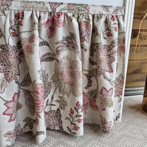 May include: A floral valance with a gathered top edge. The fabric features a large-scale pattern of pink and burgundy flowers and leaves on a neutral background. The valance is hanging in front of a white window frame.