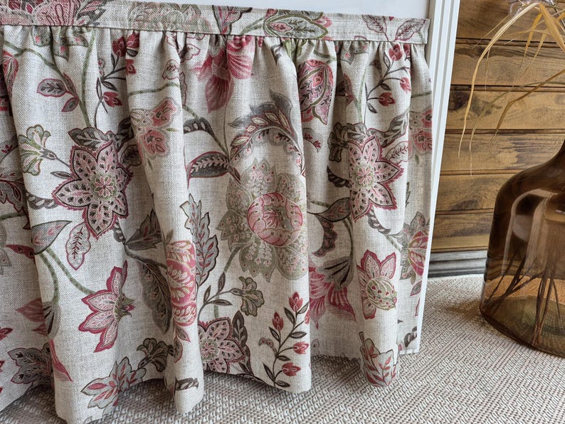 May include: A floral valance with a gathered top edge. The fabric features a large-scale pattern of pink and burgundy flowers and leaves on a neutral background. The valance is hanging in front of a white window frame.