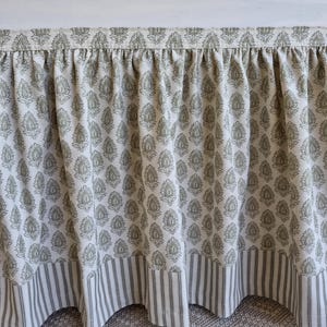 May include: A bed skirt with a floral pattern in shades of green and white. The bottom edge features a striped fabric in the same color scheme. The fabric is gathered at the top, creating a ruffled effect.