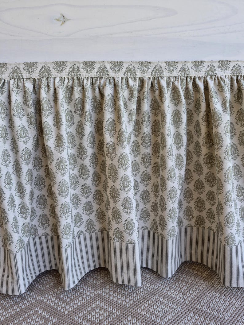 May include: A bed skirt with a floral pattern in shades of green and white. The bottom edge features a striped fabric in the same color scheme. The fabric is gathered at the top, creating a ruffled effect.