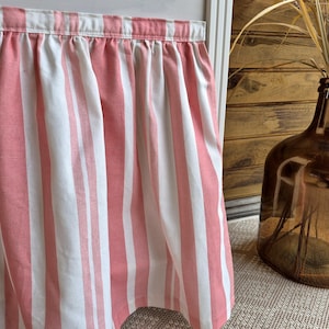 Puede incluir: Una cenefa con volantes y rayas verticales blancas y rosa coral. La tela está fruncida en la parte superior, creando pliegues suaves. Una botella de vidrio marrón con plantas secas está cerca.