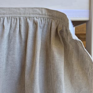 May include: A beige linen curtain panel featuring a gathered top edge with a white hook-and-loop fastener strip attached to the back. The fabric has a visible woven texture and hangs in soft, vertical folds against a white frame structure.