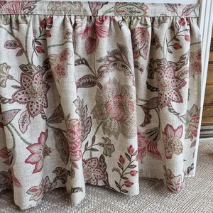 May include: Floral patterned curtain panel in shades of beige, pink, and green. The fabric features a repeating design of large flowers and leaves. The curtain is gathered at the top, creating soft folds.