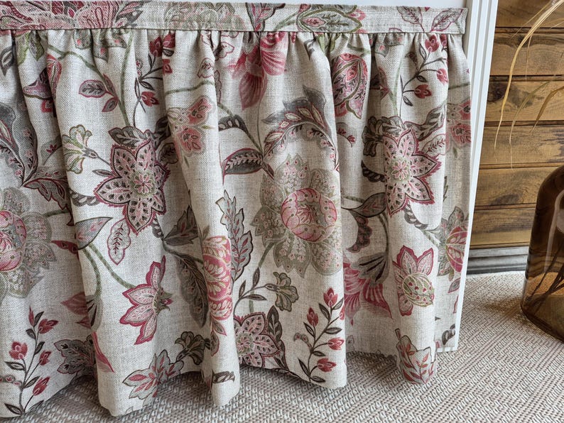 May include: Floral patterned curtain panel in shades of beige, pink, and green. The fabric features a repeating design of large flowers and leaves. The curtain is gathered at the top, creating soft folds.