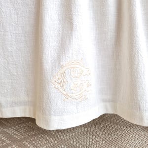 Può includere: Tessuto di lino color crema con superficie strutturata. Il tessuto è ricamato con un monogramma decorativo in una tonalità crema più chiara. Il bordo inferiore del tessuto è leggermente curvo.