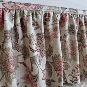 May include: Close-up of a floral valance with a gathered design. The fabric features a pattern of large pink and red flowers with green and gray leaves on a beige background. The valance is likely for a window.