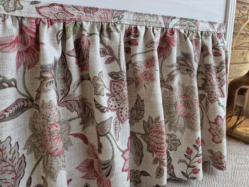 May include: Close-up of a floral valance with a gathered design. The fabric features a pattern of large pink and red flowers with green and gray leaves on a beige background. The valance is likely for a window.