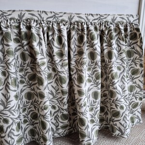 May include: A bed skirt with a gathered design, showcasing a botanical print. The fabric has a white base adorned with a repeating pattern of green and grey leaves and fruit. The skirt is intended to hang from the bed frame.