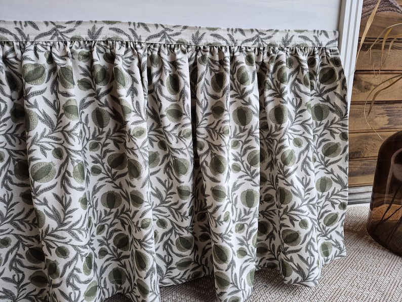 May include: A bed skirt with a gathered design, showcasing a botanical print. The fabric has a white base adorned with a repeating pattern of green and grey leaves and fruit. The skirt is intended to hang from the bed frame.