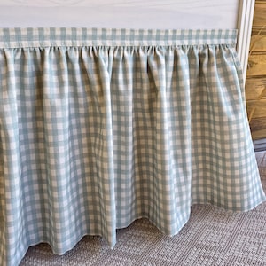 May include: A light blue and white gingham patterned bed skirt. The fabric is gathered at the top, creating a ruffled effect. The skirt hangs down, displaying the checkered pattern. The bed skirt is made of a soft, lightweight fabric.