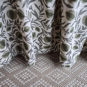 May include: Close-up of a white fabric featuring a repeating pattern of green fruit and grey leaves. The fabric is draped, showing its texture and design. The fabric rests on a textured, light brown surface.