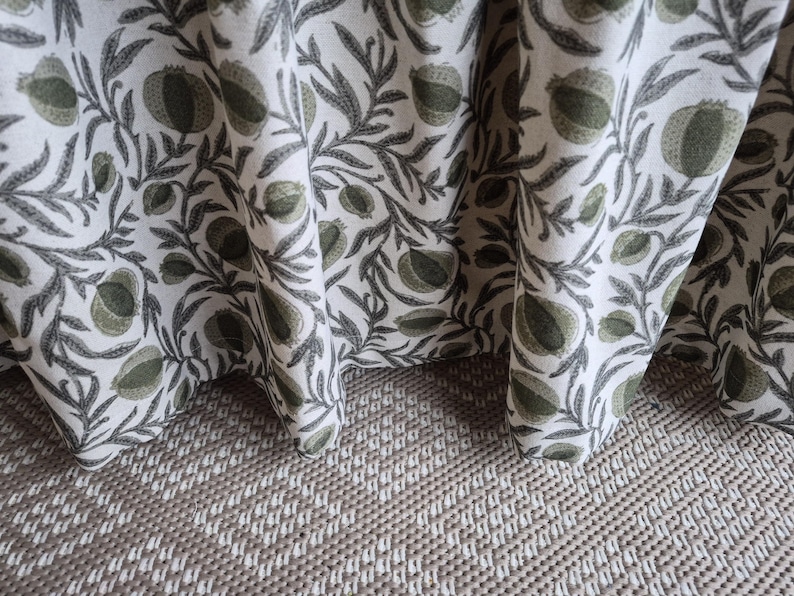 May include: Close-up of a white fabric featuring a repeating pattern of green fruit and grey leaves. The fabric is draped, showing its texture and design. The fabric rests on a textured, light brown surface.