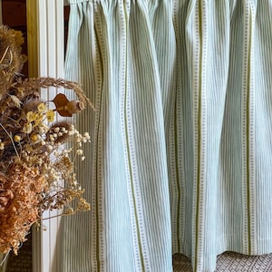 May include: A light green and white striped curtain. The curtain has vertical stripes of varying widths, with a dotted pattern in between. The fabric is gathered at the top, creating soft folds. The curtain is hanging in front of a white door frame.