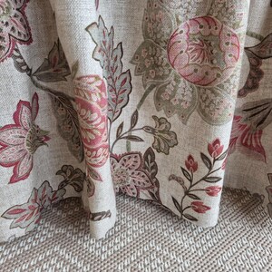 May include: Close-up of a floral fabric with a beige background. The fabric features large, stylized flowers in shades of pink, red, and olive green. The design includes detailed leaves and stems, creating a textured, botanical pattern. The fabric is draped, showing its folds and texture.